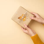 Top view photo of hands in yellow sweater giving craft paper gift box with twine bow and yellow autumn leaf.