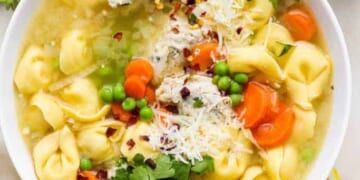 A bowl of tortellini soup with carrots, peas, and garnished with parsley and grated cheese.