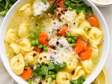 A bowl of tortellini soup with carrots, peas, and garnished with parsley and grated cheese.