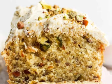A close-up image of a sliced pistachio cake with creamy frosting and crushed nuts on top.