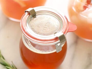 A glass jar with orange juice, honey simple syrup, and a rosemary sprig.