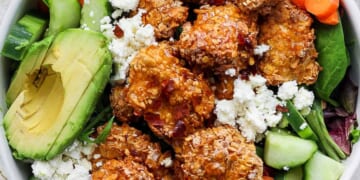 A bowl containing fried chicken, avocado slices, cherry tomatoes, sliced cucumbers, carrots, and leafy greens, topped with crumbled cheese.