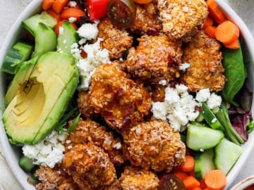 A bowl containing fried chicken, avocado slices, cherry tomatoes, sliced cucumbers, carrots, and leafy greens, topped with crumbled cheese.