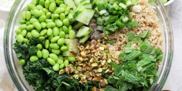 A bowl of salad containing quinoa, kale, chopped cucumbers, green onions, shelled edamame, chopped nuts, and fresh herbs, with a pair of golden spoons on the.