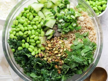 A bowl of salad containing quinoa, kale, chopped cucumbers, green onions, shelled edamame, chopped nuts, and fresh herbs, with a pair of golden spoons on the.